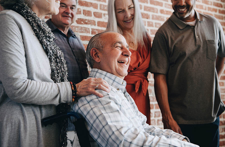 A happy family surrounds an elderly man in a wheelchair showing they are happy he is at home rather than a residential carehome