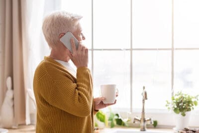 Elderly Lady Looking Out The Window Holding A Mug In One Hand, And Holding Her Phone In The Other