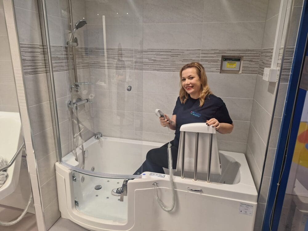 Sarah sat on an seat lift in an accessible walk in bath with glass door at the mobility bathroom showroom in Henley-on-Thames.