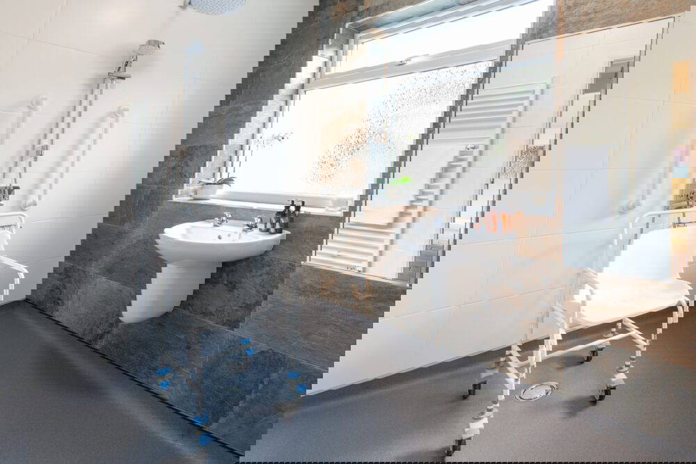 Wheelchair accessible wetroom with a mobile shower chair, wall-mounted grab rails, walk-in shower area and modern sink beside a large frosted window.